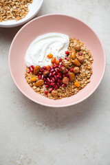 Roseate bowl with granola, greek yogurt, pomegranate and nuts, vertical shot on a light-beige stone background, elevated view