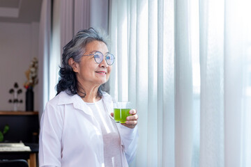 Senior Asian lady drinking glass of herbal supplement drink while looking out the window with copy space for nutrition boosting and healthy organics product consumption