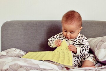 Fototapeta premium A cute little baby boy is carefully looking at a book with interest