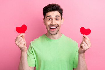 Photo portrait of young astonished man wearing green t shirt holding red paper postcards heart figures isolated on pink color background