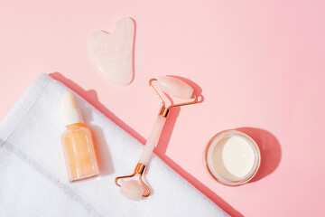 Cosmetic serum bottle, cream jar and face roller, gua sha stone on pink background in sunlight. Skin care, spa and wellness concept. Top view, flat lay