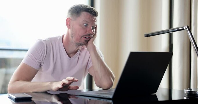 Bored man checking online laptop content sitting on table at home. Nervous emotions of expectation and disappointment