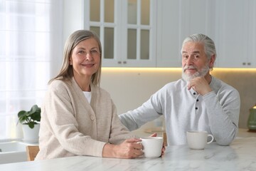 Affectionate senior couple with cups of drink at white marble table in kitchen