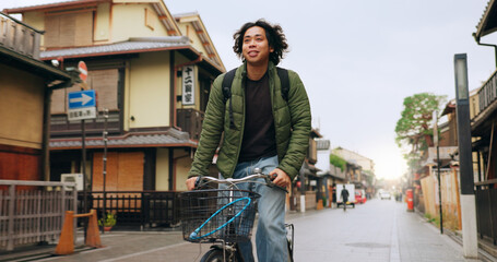 Bicycle, cycling and man by buildings for travel, exercise or transportation to college academy. Fitness, backpack and young male student driving a bike to university for carbon footprint by houses. - obrazy, fototapety, plakaty