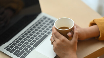 Cropped shot young woman holding coffee cup and using laptop on wooden desk
