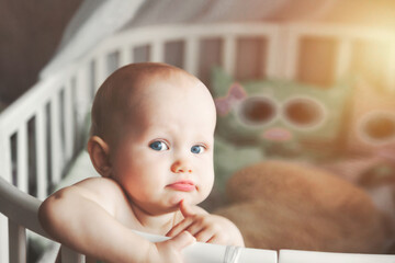 Thoughtful serious little kid looking at camera from infant cot at nursery. Adorable hairless baby...