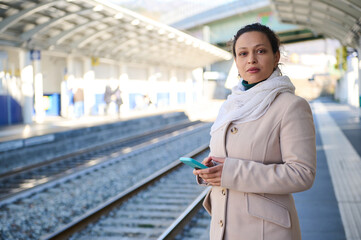 Pensive young woman waiting for high-speed train, holding mobile phone, standing on the platform of...