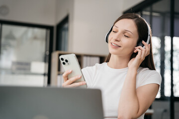 Smiling girl relaxing at home, she is playing music using smartphone tablet, laptop, and wearing...