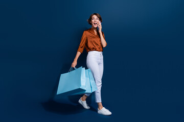 Full length photo of excited dreamy girl dressed brown shirt holding shoppers talking device empty space isolated blue color background