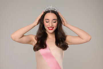 Beautiful young woman with tiara and ribbon in dress on grey background. Beauty contest