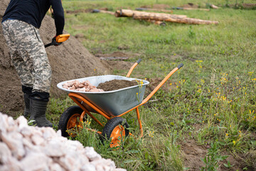 A man is loading crushed stones into the wheelbarrow with a shovel. Laying the foundation, building...