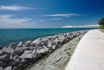 View of the blue sky with white clouds far in the background, the turquoise sea and the contrasting white, wide empty promenade in the tourist town of Grado, Italy.