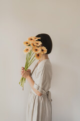 Beautiful woman in linen dress holding pastel gerber flowers bouquet over white wall. Aesthetic...