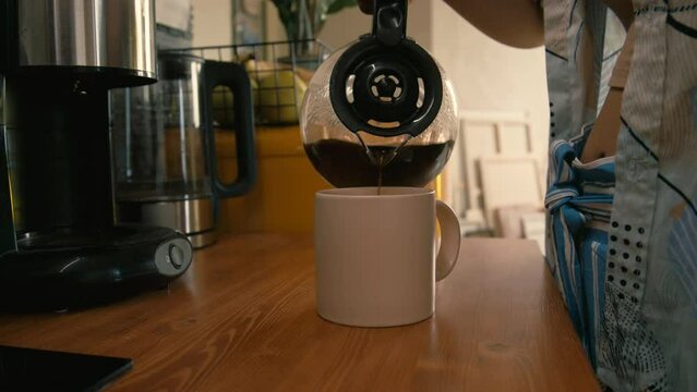Side shot of unrecognizable lady pouring coffee from coffeemaker into mug and going away at home