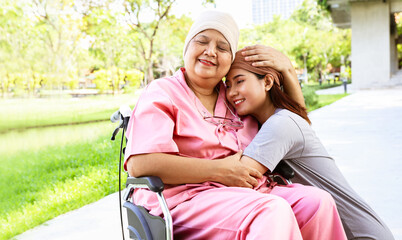 Portrait senior mother with cancer wearing headscarf sits in a wheelchair with her cute daughter wearing headscarf standing together lovingly embracing her mother encouraging her to wish her well. - obrazy, fototapety, plakaty