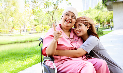 Naklejka premium Portrait senior mother with cancer wearing headscarf sits in a wheelchair with her cute daughter wearing headscarf standing together lovingly embracing her mother encouraging her to wish her well.