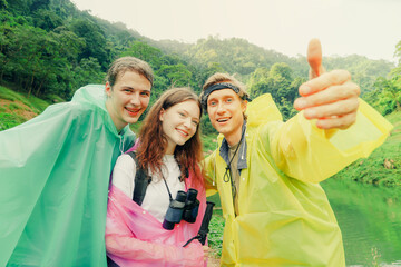 Portrait Close friends, handsome man and beautiful caucasian woman travel hiking in the rainy season and wear raincoats together taking hilarious selfies looking joyfully at the camera.