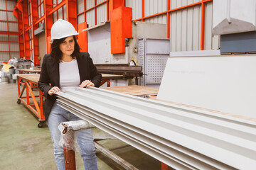 Obraz premium Female manager engineer wearing safety helmet inspects the production quality of white coated rolled metal roofing sheets, in a standardized and safe warehouse production facility.