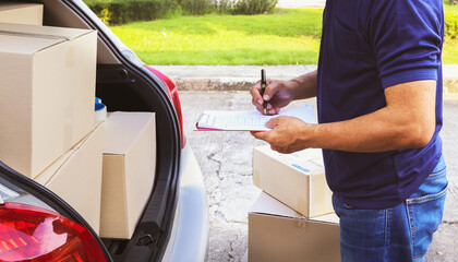 Home delivery business : Senior male employee stands at the back of the truck, recording the customer's parcel delivery list and cardboard box products on a clipboard for accurate delivery.