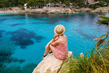 A woman in straw hat sitting on a cliff, looking towards Cala Gat beach in Mallorca