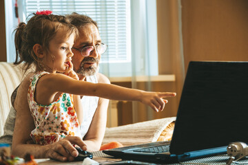 A little girl and her grandfather using a laptop together. Two generations learning how to use technologies. - obrazy, fototapety, plakaty