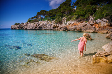 A woman in a summer dress and straw hat enjoying a stroll in the water along Cala Gat beach in...