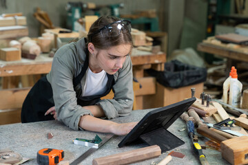 A young woman is training to be a carpenter in the workshop. smartphone and working with a laptop computer in a wood workshop. Black female carpenters contact customers by smartphone. SME orders. - obrazy, fototapety, plakaty