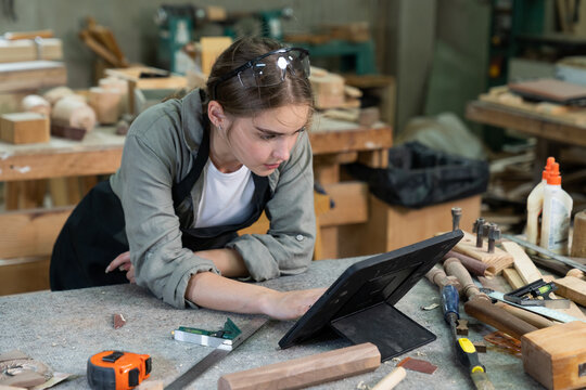 A young woman is training to be a carpenter in the workshop. smartphone and working with a laptop computer in a wood workshop. Black female carpenters contact customers by smartphone. SME orders.