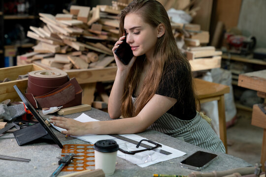 A young woman is training to be a carpenter in the workshop. smartphone and working with a laptop computer in a wood workshop. Black female carpenters contact customers by smartphone. SME orders.