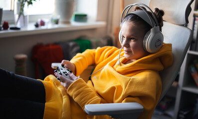 Teenage girl with headset is sitting in a comfortable computer chair, holding a white gamepad in her hands and playing a game on a PC or console. 