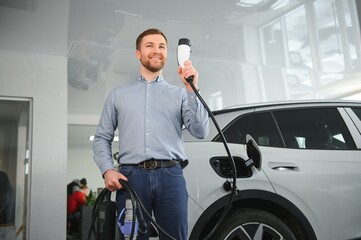 Handsome business man holding charging cable for electric car. Caucasian male stands near electric auto in dealership © Serhii