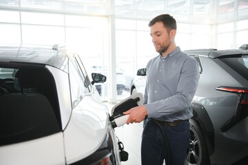 Handsome business man holding charging cable for electric car. Caucasian male stands near electric...