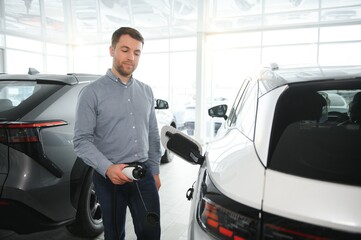 Beard man trying a new charging cable with a car charging station at the motor dealership. Concept of buying electric vehicle. Smart ecological living