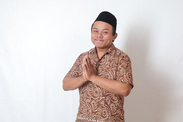 Portrait of smiling Asian man wearing batik shirt and songkok showing apologize and welcome hand gesture. Ramadan advertising concept. Isolated image on gray background