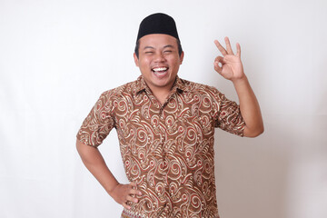 Portrait of excited Asian man wearing batik shirt and songkok showing ok hand gesture and smiling...