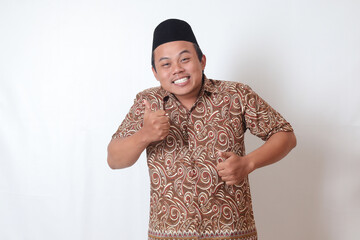 Portrait of excited Asian man wearing batik shirt and songkok smiling and looking at camera, making...