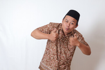 Portrait of excited Asian man wearing batik shirt and songkok smiling and looking at camera, making...