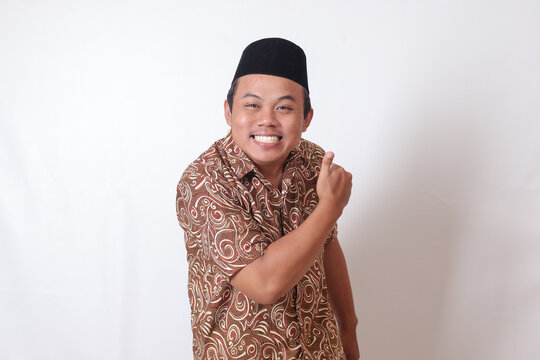 Portrait of excited Asian man wearing batik shirt and songkok smiling and looking at camera, making thumbs up hand gesture. Isolated image on gray background