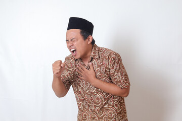 Portrait of unwell Asian man wearing batik shirt and songkok keeping hand palm covering his mouth...