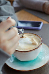 A cup of cappuccino coffee and a girl's hand stirring the foam from the coffee with a spoon