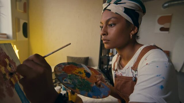 Side chest up of female left-handed Black artist painting pink and yellow flowers on canvas using paintbrush and palette with vivid paint in modern house