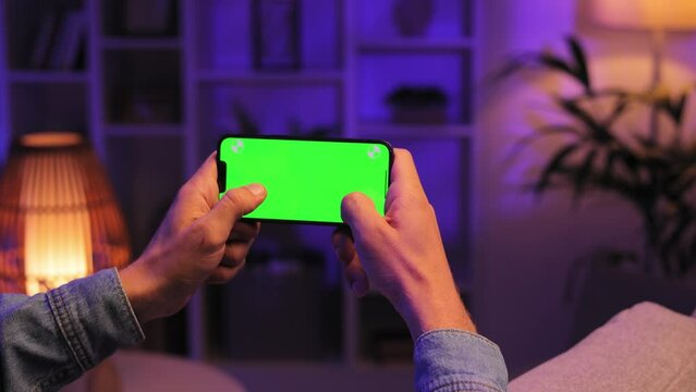 Closeup shot of a man sitting on a sofa using a smartphone in a horizontal position with green screen mockup in a evening room, use two fingers for gaming.