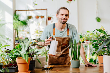 Handsome man wearing apron carefully watering potted houseplants indoors. Concept of plants care and home garden.