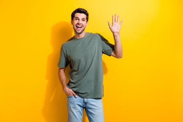Photo of optimistic man with bristle dressed khaki t-shirt waving palm say hi hand in pocket isolated on vibrant yellow color background © deagreez
