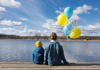 woman and a child sit with their backs near a lake on a sunny day, holding yellow and blue balloons...