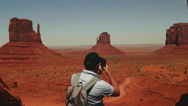Back view young adult tourist man with backpack takes smartphone photo of epic desert view at Monuments Valley, Arizona.