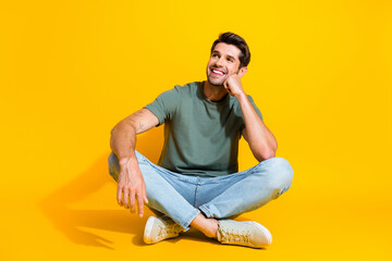 Full length photo of positive dreamy man dressed khaki t-shirt sitting legs crossed looking empty...