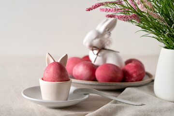Easter holiday table decor, red colored egg in cup on saucer, spoon, on neutral beige linen tablecloth, blurred plate with eggs and bunny figurine, spring flowers in vase on background