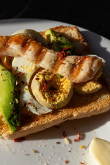 A healthy breakfast of toast topped with boiled egg, avocado, and grilled chicken breast, served on a white plate bathed in sunlight from a window.