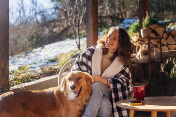 Young adult happy woman cuddling with her dog on the terrace of a country house in early spring - obrazy, fototapety, plakaty
